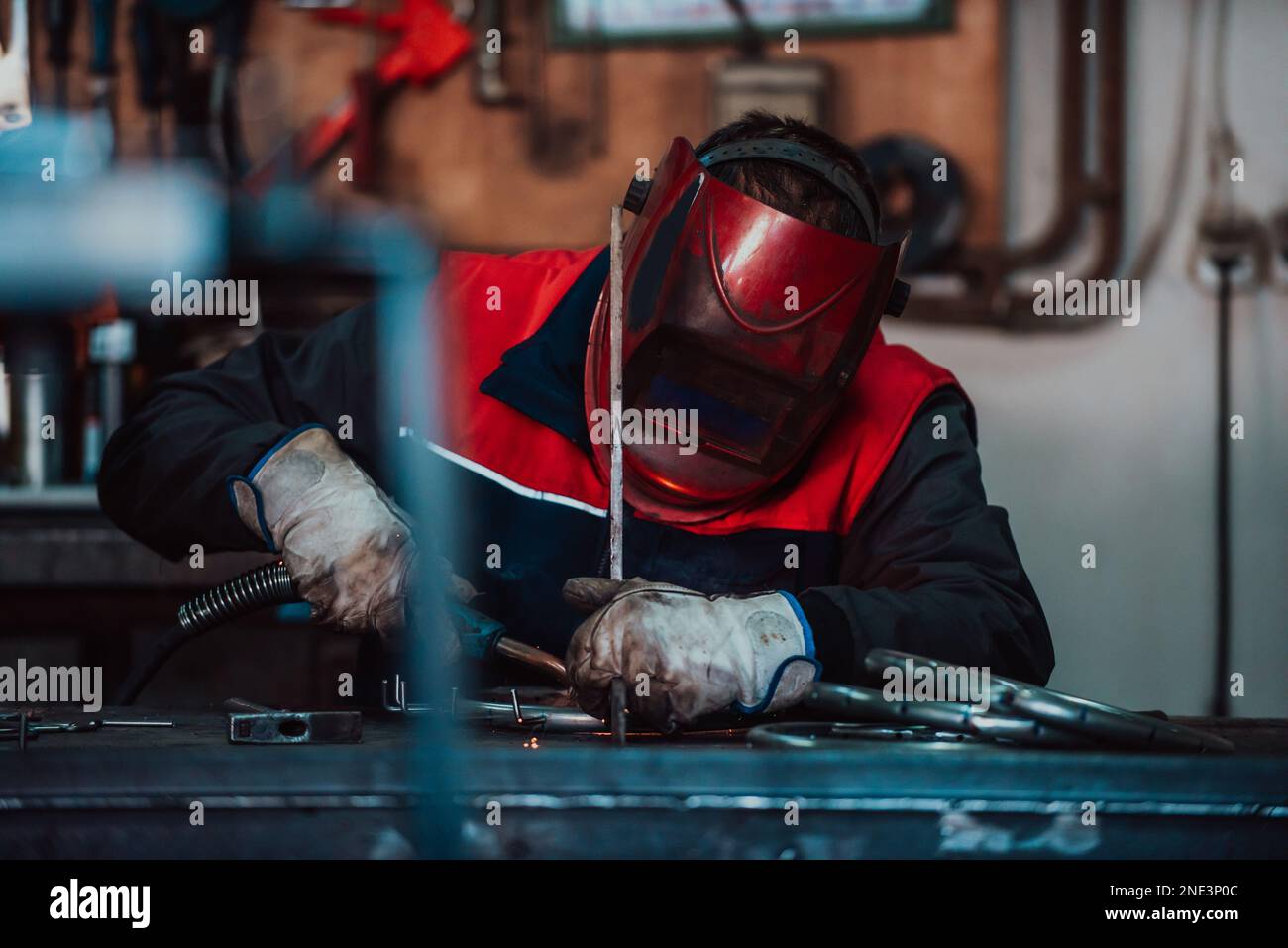 Saldatrice professionale in uniforme protettiva e maschera saldante tubo metallico sul tavolo industriale con altri lavoratori dietro nell'officina industriale. Foto Stock