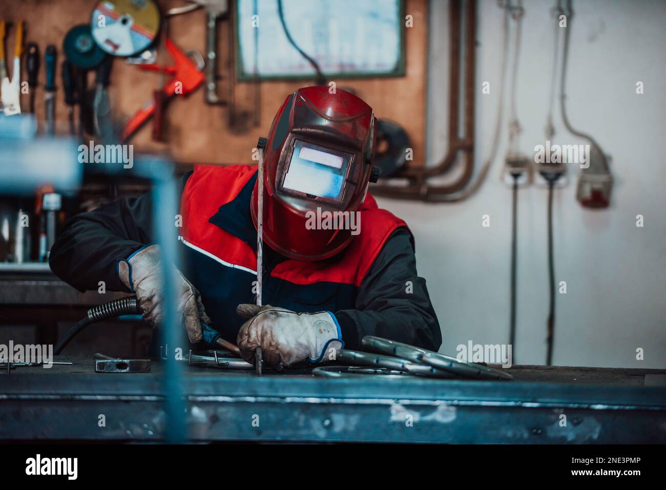Saldatrice professionale in uniforme protettiva e maschera saldante tubo metallico sul tavolo industriale con altri lavoratori dietro nell'officina industriale. Foto Stock
