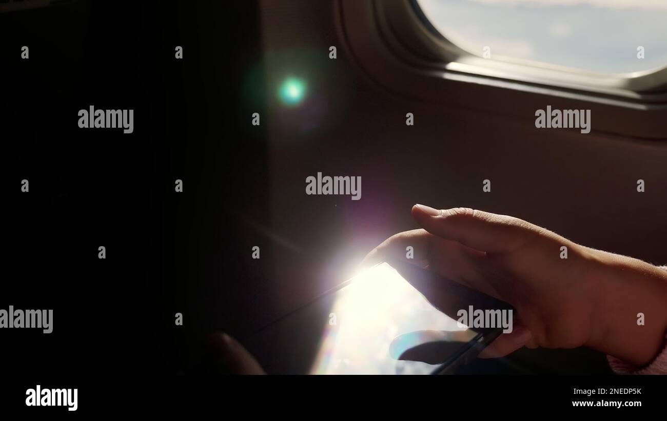 primo piano. i raggi del sole si riflettono nel telefono. sagoma scura delle mani dei bambini e del telefono cellulare contro l'illuminatore dell'aeroplano. Bambini che usano, gioca su smartphone, tablet nella cabina dell'aereo. Foto di alta qualità Foto Stock