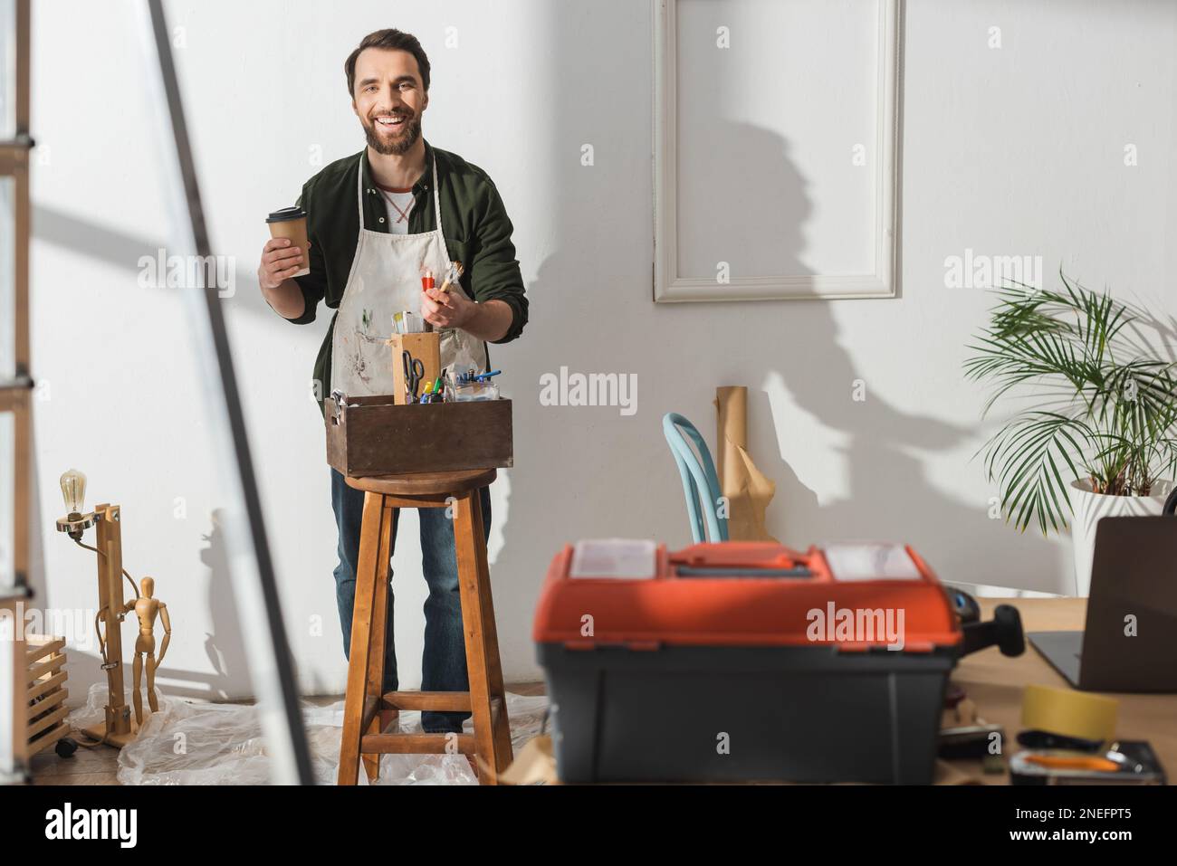 Sorridente artigiano che tiene il caffè da andare e pennelli in officina, immagine stock Foto Stock