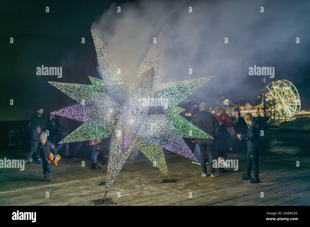 Southend on Sea, Regno Unito. 16 febbraio 2023. Scena notturna sul Southend Pier con una grande scultura illuminata a forma di stella circondata dai visitatori. L'installazione si illumina di colori diversi, verde, viola, bianco, in mezzo alla nebbia e all'illuminazione festosa. Una ruota panoramica e giostre sullo sfondo evocano festeggiamenti stagionali e il coinvolgimento della comunità. Il Luminocity Light Festival torna a Southend per un secondo anno. Penelope Barritt/Alamy Live News Foto Stock