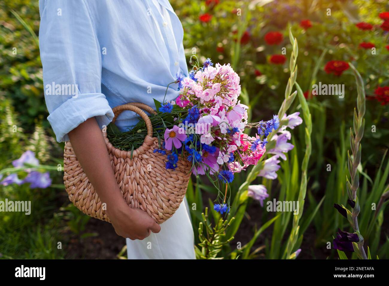Primo piano del sacchetto di paglia riempito con fiori blu e rosa. Donna in possesso di borsa estiva con bouquet di fiori. Accessori femminili alla moda Foto Stock