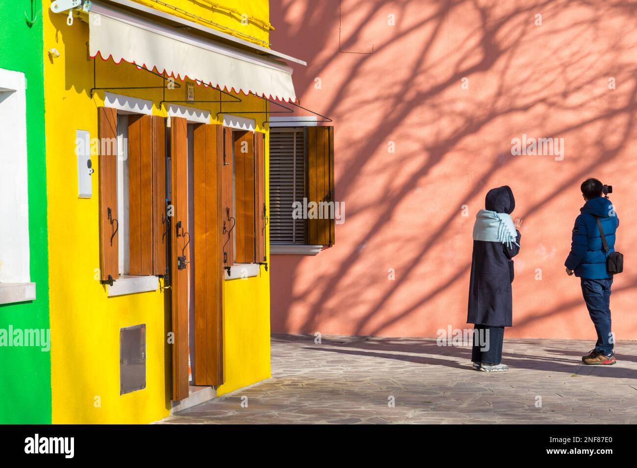 Due turisti che guardano le case dai colori luminosi e le ombre degli alberi sul lato dell'edificio a Burano, Venezia, Italia nel mese di febbraio Foto Stock