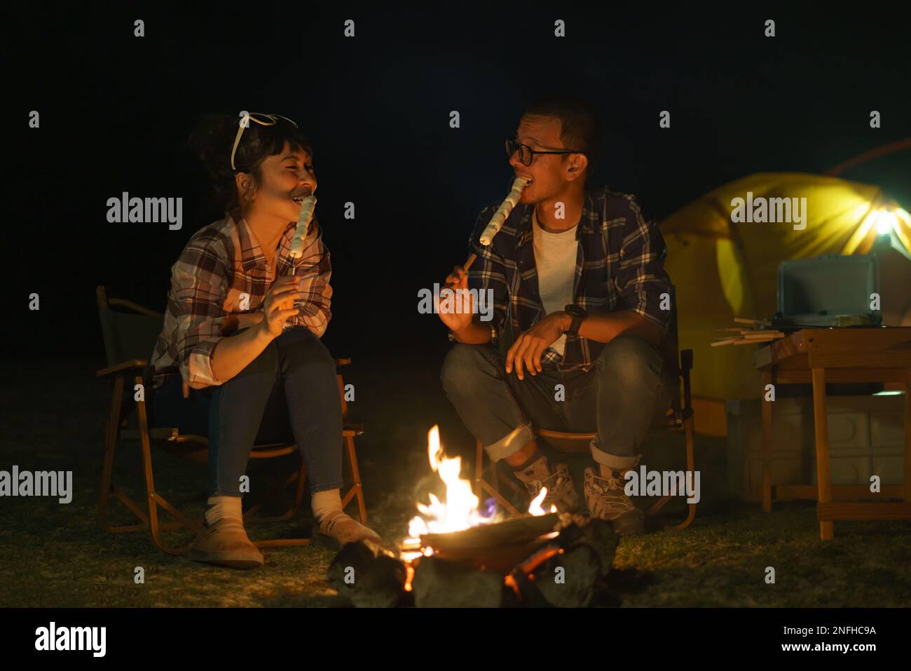 Coppie asiatiche arrostire insieme marshmallows ad un falò dove hanno messo su una tenda per accampare vicino al lago di notte. Foto Stock