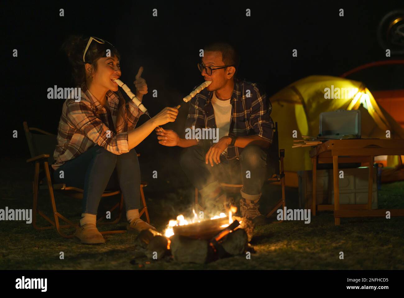 Coppie asiatiche arrostire insieme marshmallows ad un falò dove hanno messo su una tenda per accampare vicino al lago di notte. Foto Stock