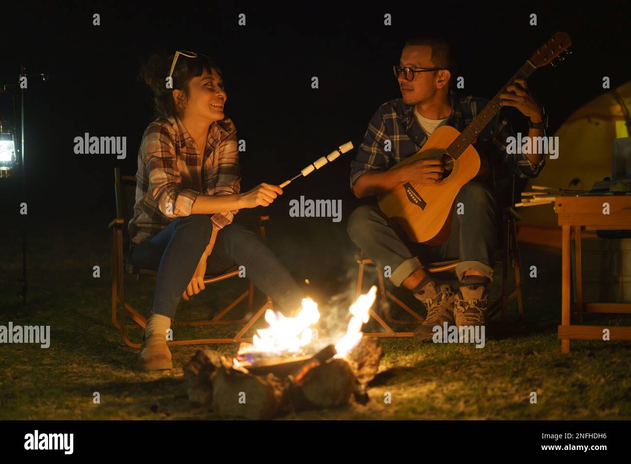Donna asiatica che brinda marshmallows mentre il suo ragazzo suona la chitarra al fuoco dove hanno messo le loro tende per accampare presso il lago di notte. Foto Stock