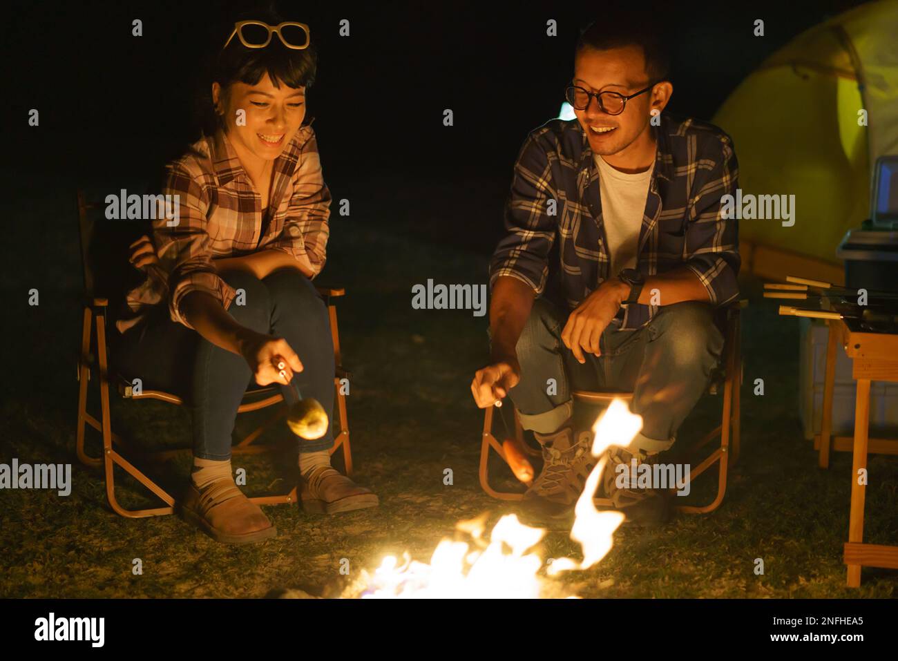 La coppia asiatica sta grigliando il mais e le salsicce in un falò dove hanno allestito una tenda per accamparsi al lago di notte. Foto Stock
