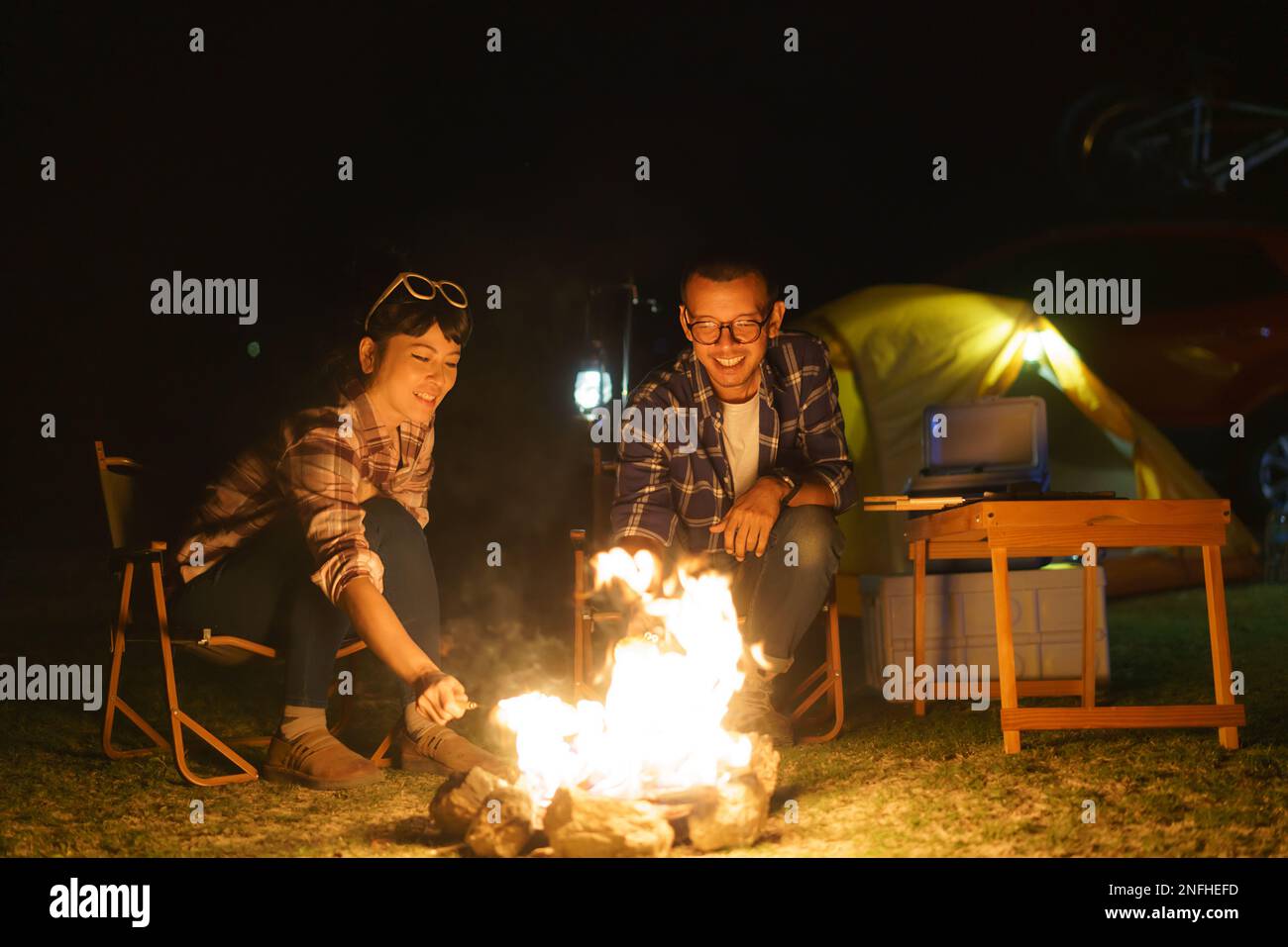Asian coppia griglia mais su un fuoco hanno impostato per illuminare la notte, mentre nel mezzo di una tenda campeggio vicino a un lago. Foto Stock
