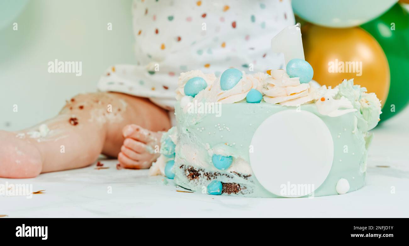 primo piano di una bella torta blu e di un bambino durante una festa di smash torta per il loro primo compleanno, e alcuni palloncini di colori diversi sullo sfondo Foto Stock