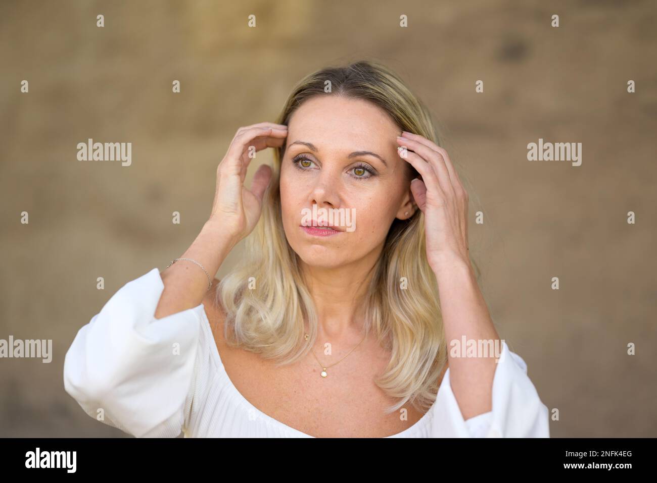 Il ritratto di una donna bionda di mezza età con una blusa bianca corre con entrambe le mani attraverso i capelli con un'espressione facciale premurosa davanti ad una fronte Foto Stock
