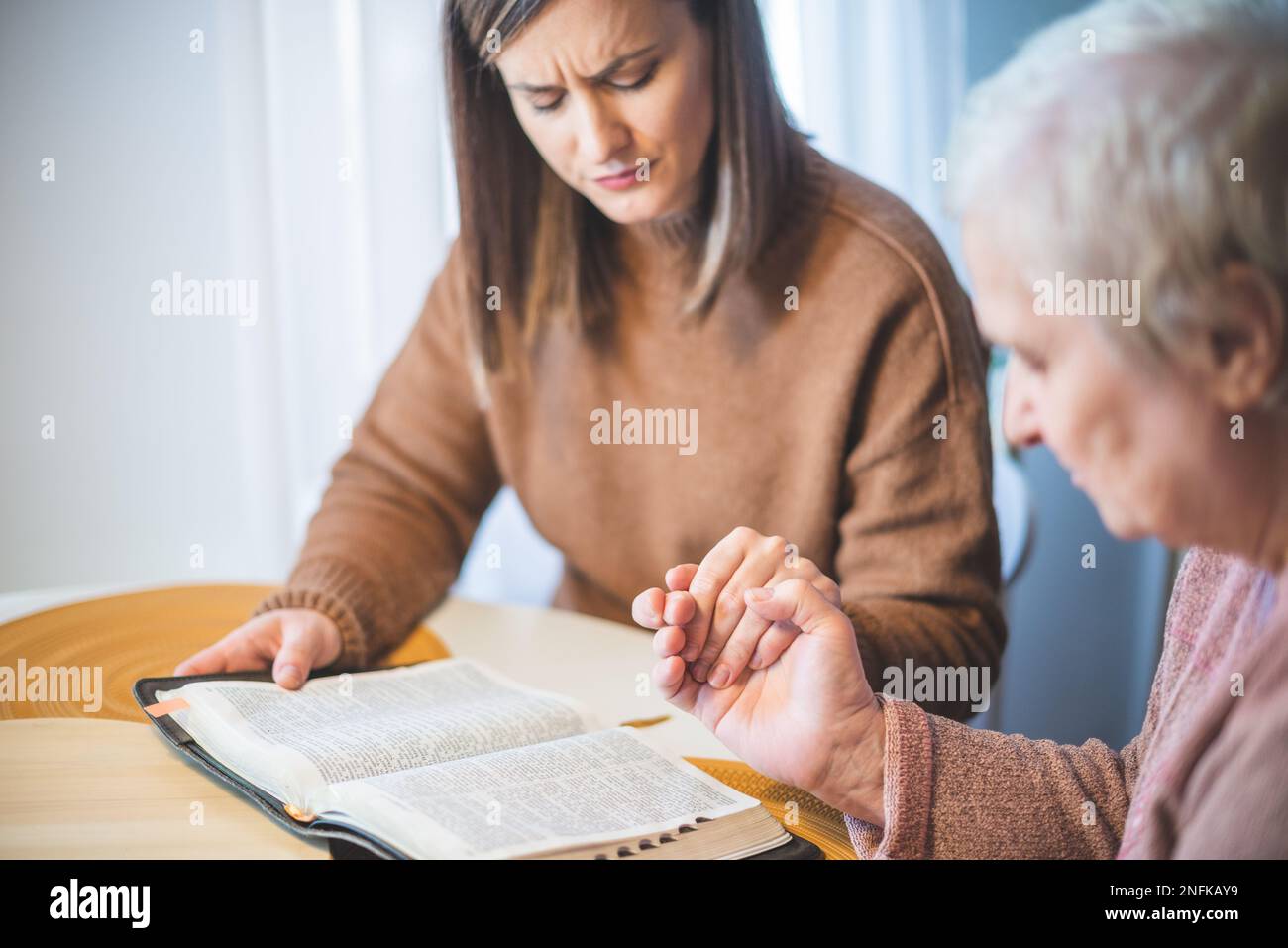 Donna anziana con la figlia che tiene le mani insieme e prega al tavolo Foto Stock