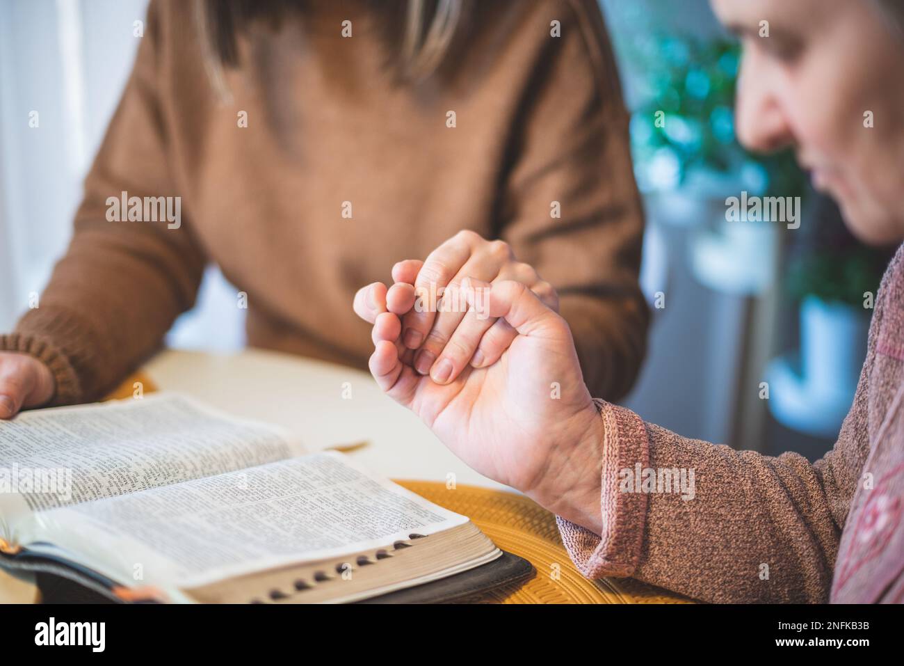 Donna anziana con la figlia che tiene le mani insieme e prega al tavolo Foto Stock