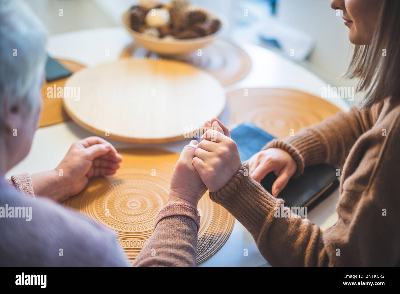 Donna anziana con la figlia che tiene le mani insieme e prega al tavolo Foto Stock
