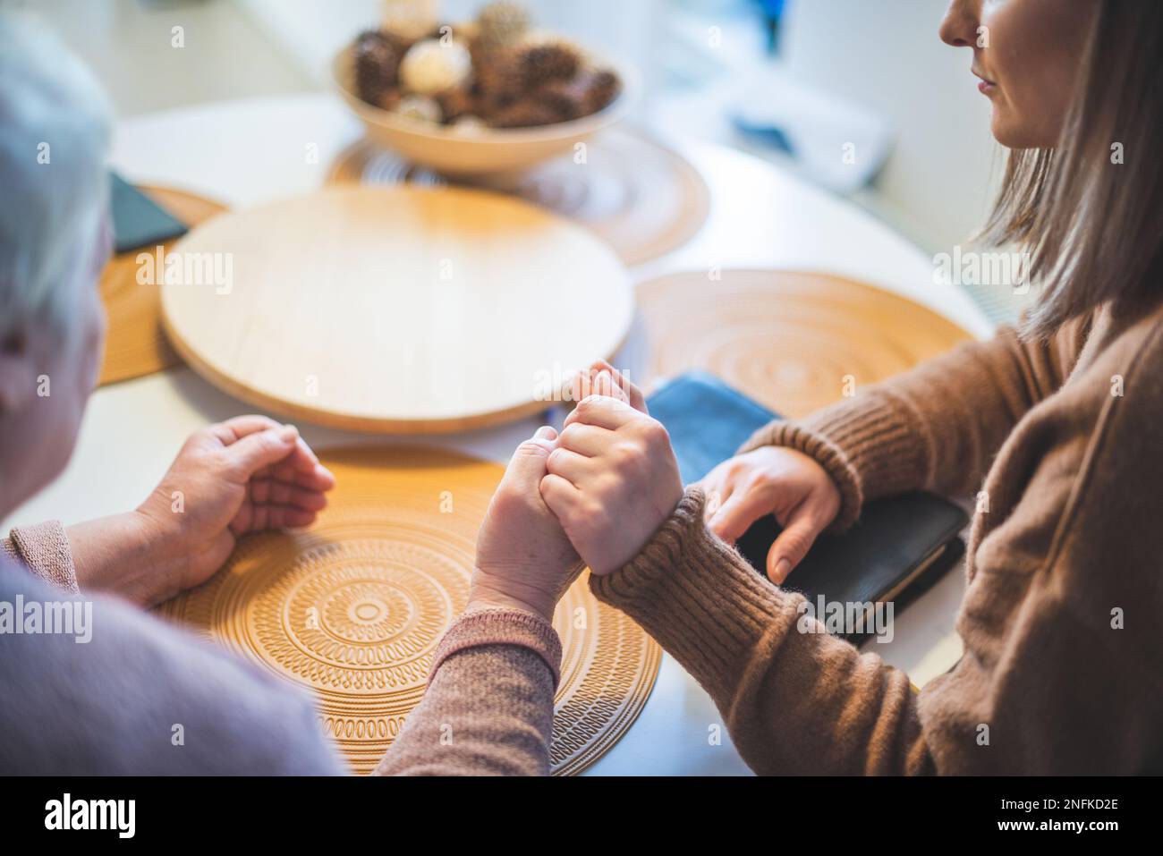 Donna anziana con la figlia che tiene le mani insieme e prega al tavolo Foto Stock