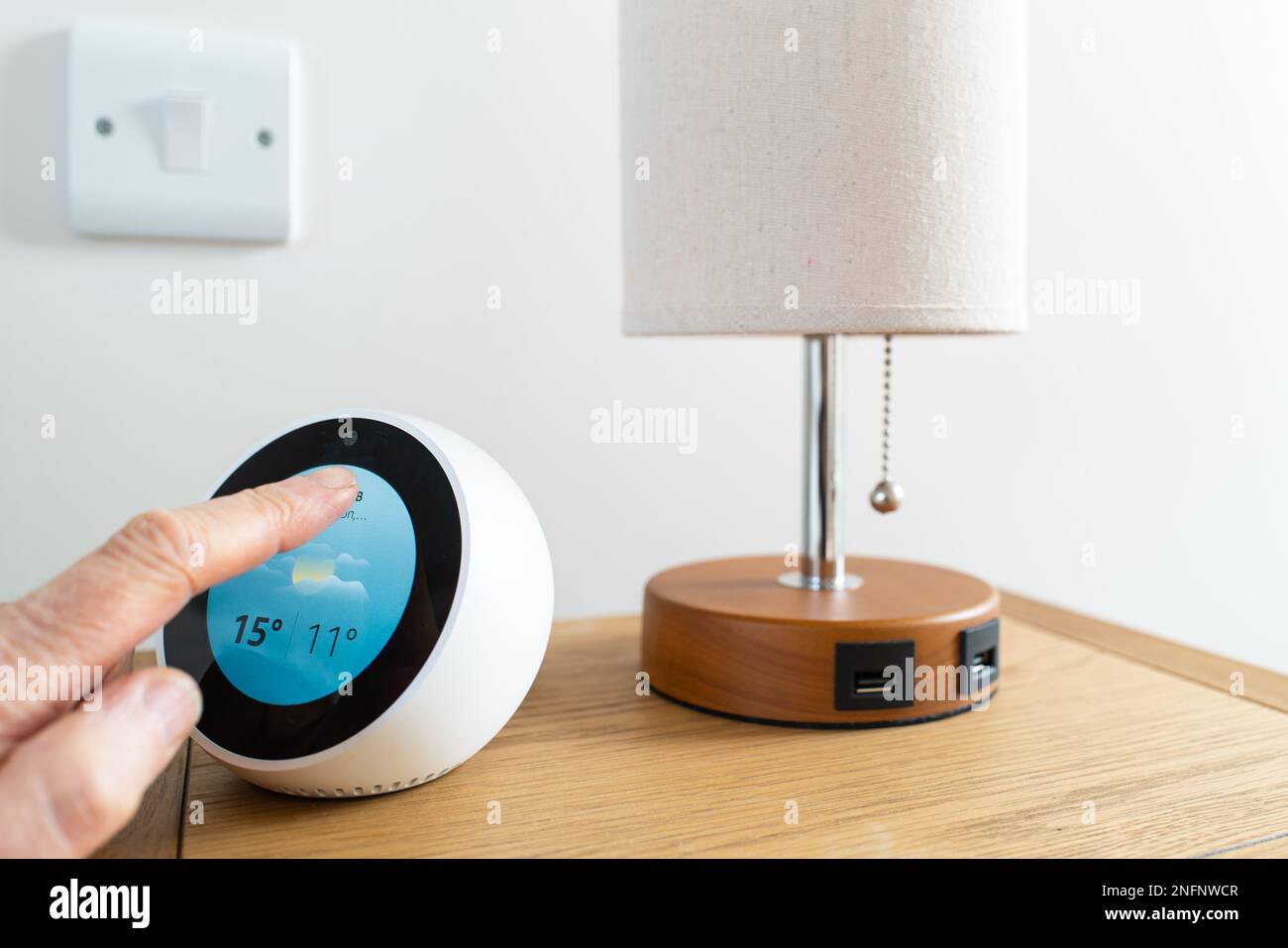 Il proprietario controlla le previsioni meteo sul suo dispositivo smart situato su un comodino. Notare la lampada da tavolo con porte di ricarica USB. Foto Stock