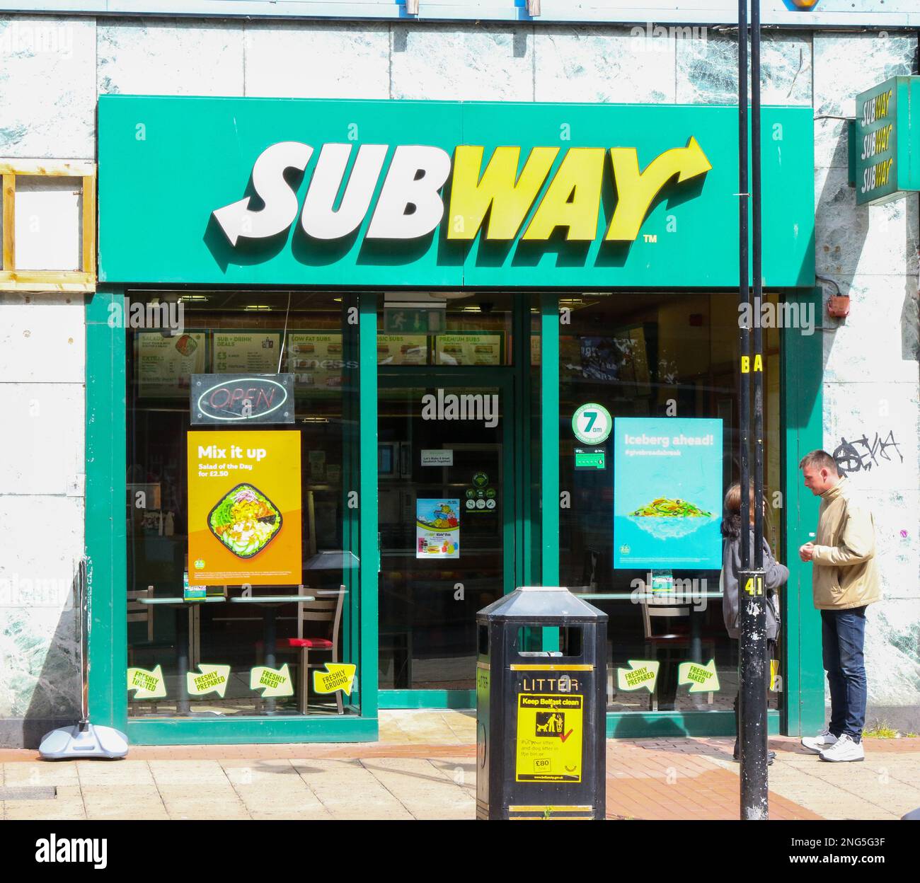 Due persone in piedi fuori da un negozio outlet in franchising Subway con cartello Subway nel centro della città del Regno Unito nelle soleggiate giornate estive. Foto Stock