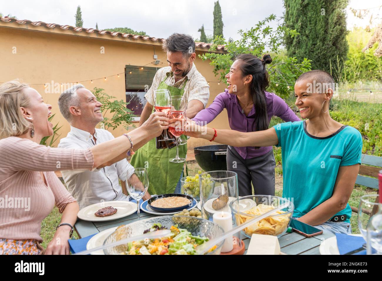 Gruppo di allegri amici di mezza età che brindano con un bel sorriso intorno al tavolo al patio della casa. Foto Stock