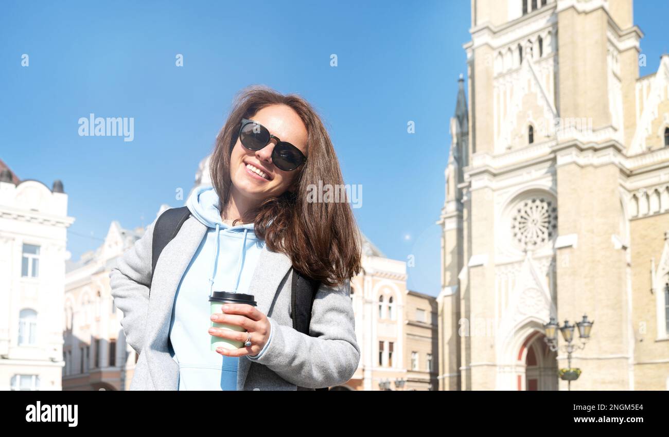 Felice giovane donna con zaino che tiene in mano la tazza di caffè mentre cammina per la città. Foto Stock