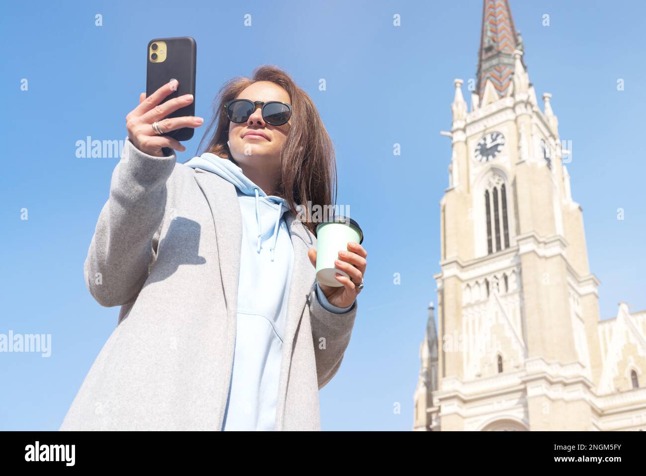Donna con occhiali da sole che indossa un cappotto in piedi sulla piazza della città vecchia tenendo in mano una tazza di caffè e un telefono cellulare. Foto Stock