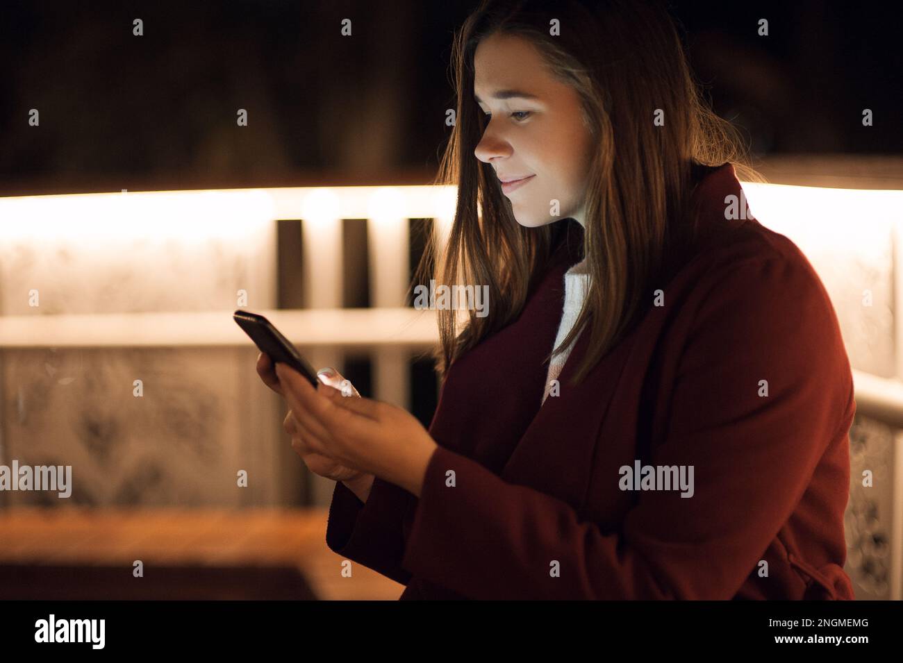Primo piano donna urbana felice e sorridente che usa uno smartphone moderno nel quartiere del centro, contro gli edifici illuminati della città Foto Stock