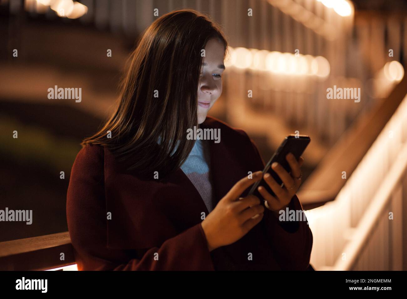 Primo piano donna urbana felice e sorridente che usa uno smartphone moderno nel quartiere del centro, contro gli edifici illuminati della città Foto Stock