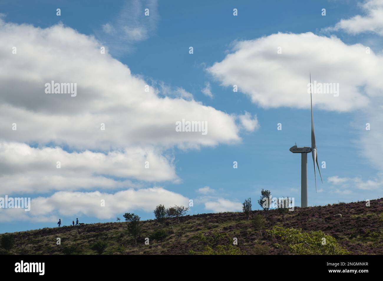 Paesaggio di brughiera con figure all'orizzonte in forma di silhouette contro un cielo blu con alcune nuvole e un'unica turbina eolica su un lato Foto Stock