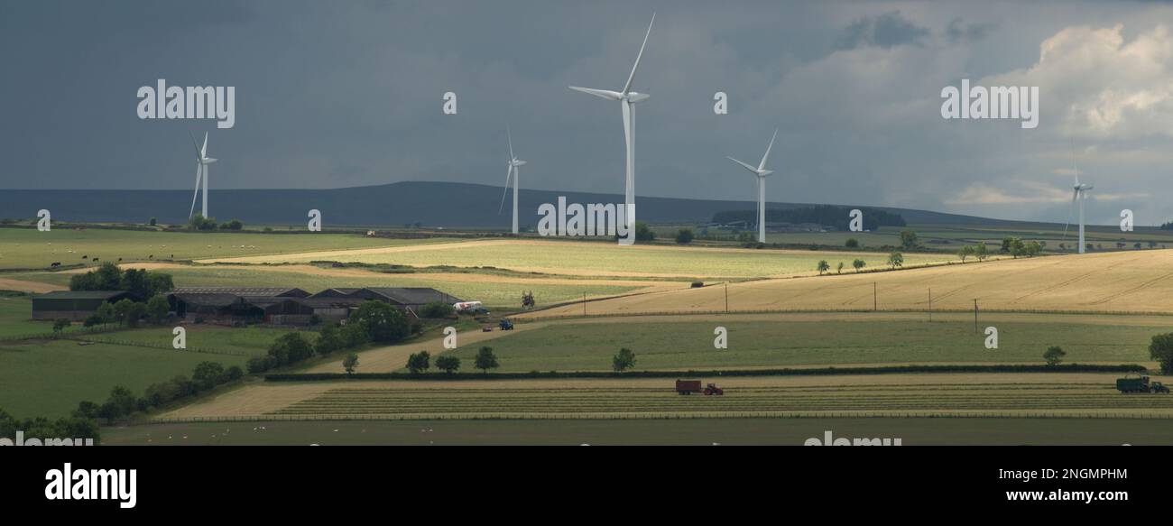 Paesaggio all'inizio dell'estate con sole che si infrange attraverso le nuvole scure ed evidenzia i campi con trattori in funzione e turbine eoliche a distanza ravvicinata Foto Stock