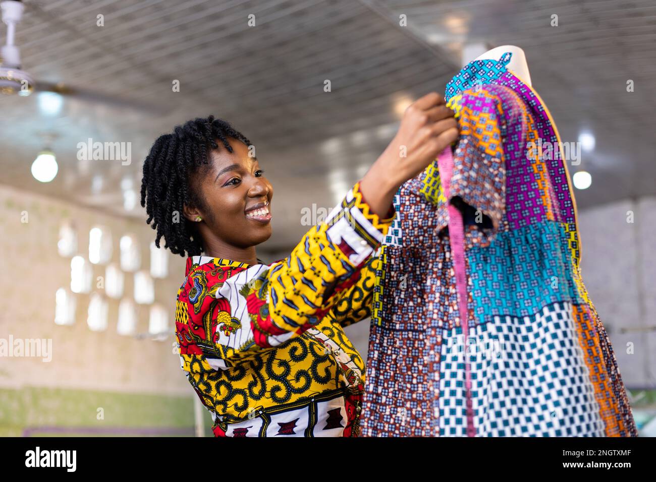 Sorridente dresssaker dell'Africa occidentale con i capelli di locs misura una parte di un vestito cucito appeso ad un manichino nel suo studio di disegno. Foto Stock