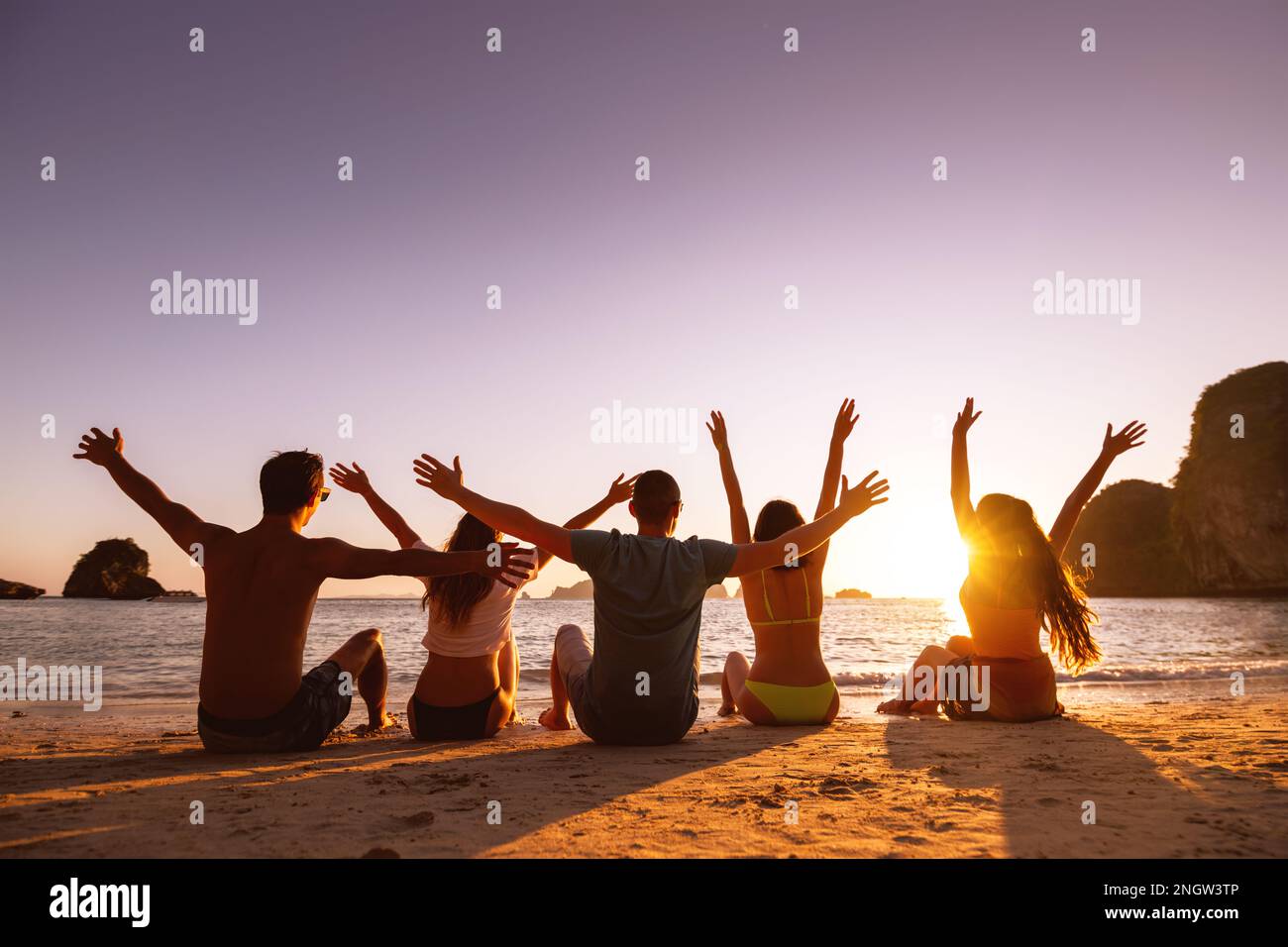 Un gruppo di giovani amici felici si diverte e si siede sulla sabbia al tramonto Foto Stock