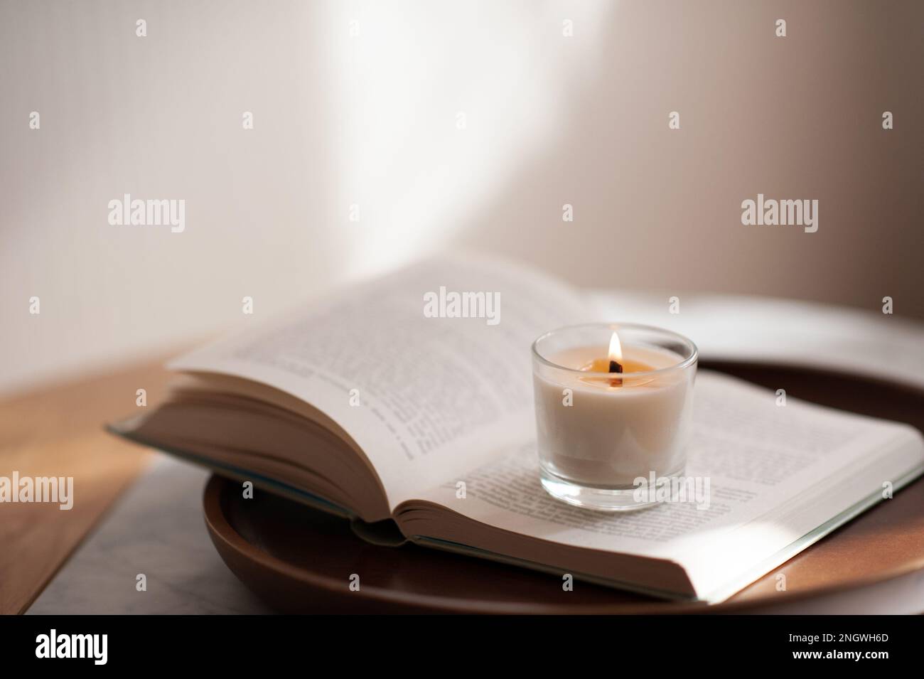 Candela bruciante profumata che rimane sul libro di carta aperto sul vassoio di legno sul closeup della tabella in soggiorno. Foto Stock