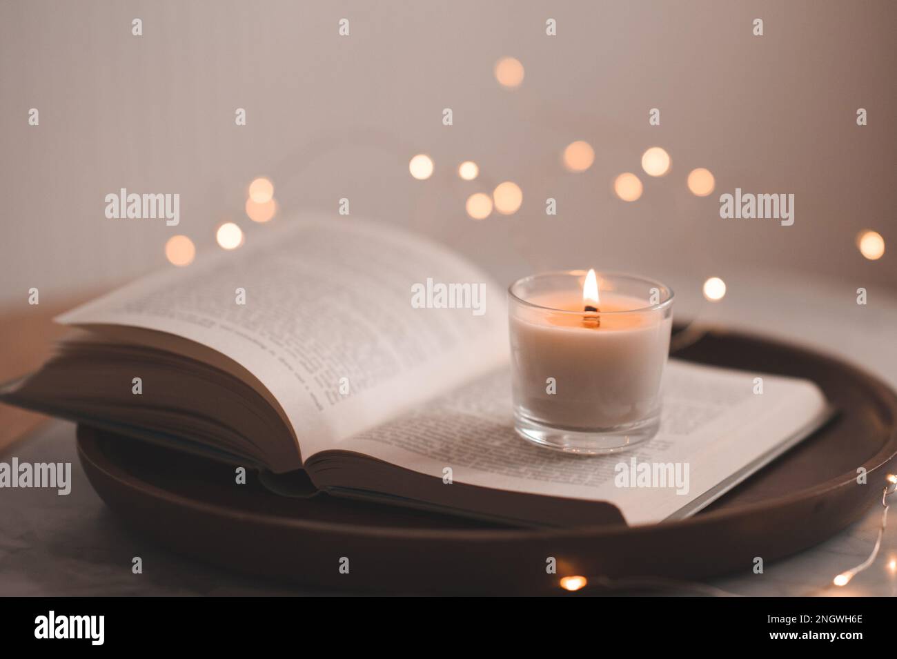 Candela bruciante profumata che rimane sul libro di carta aperto sul vassoio di legno sopra le luci di incandescenza sul tavolo nella camera da letto. Atmosfera accogliente casa. Foto Stock