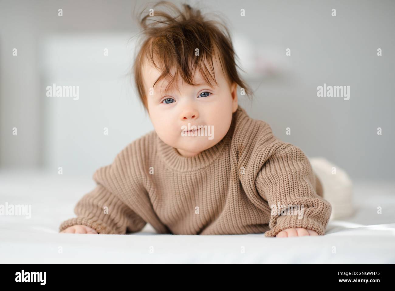 Cute ragazza neonato divertente sotto i 1 anni con capelli biondi svegliarsi indossare maglia beige maglione strisciante a letto in camera da letto closeup. Guarda la fotocamera. Foto Stock