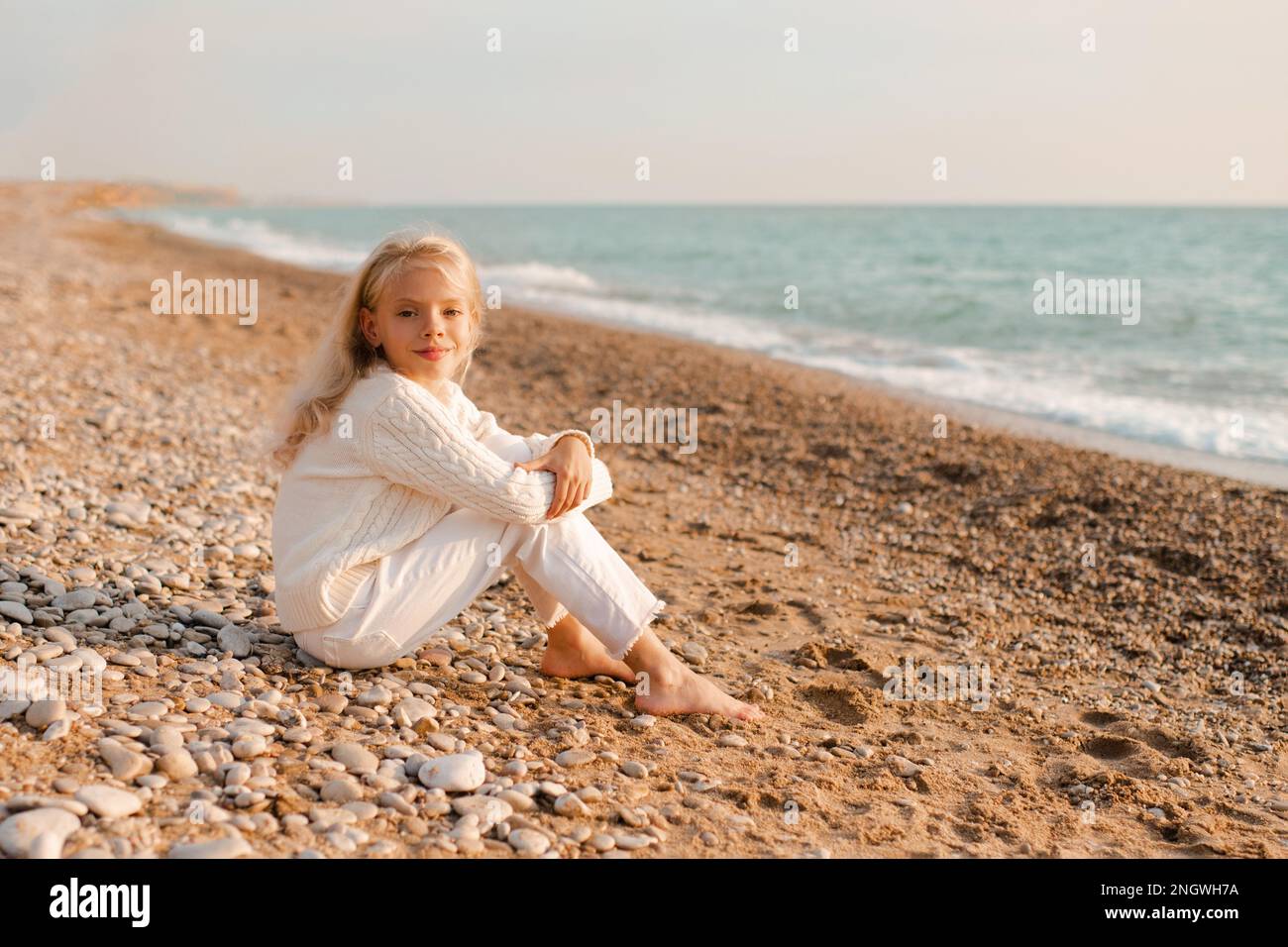 Sorridente ragazza cute capretto 6-7 anni indossare casual maglia jumper e pantaloni bianchi jeans seduti sulla sabbia sulla spiaggia sul mare riva all'aperto. Guardando la fotocamera. S Foto Stock
