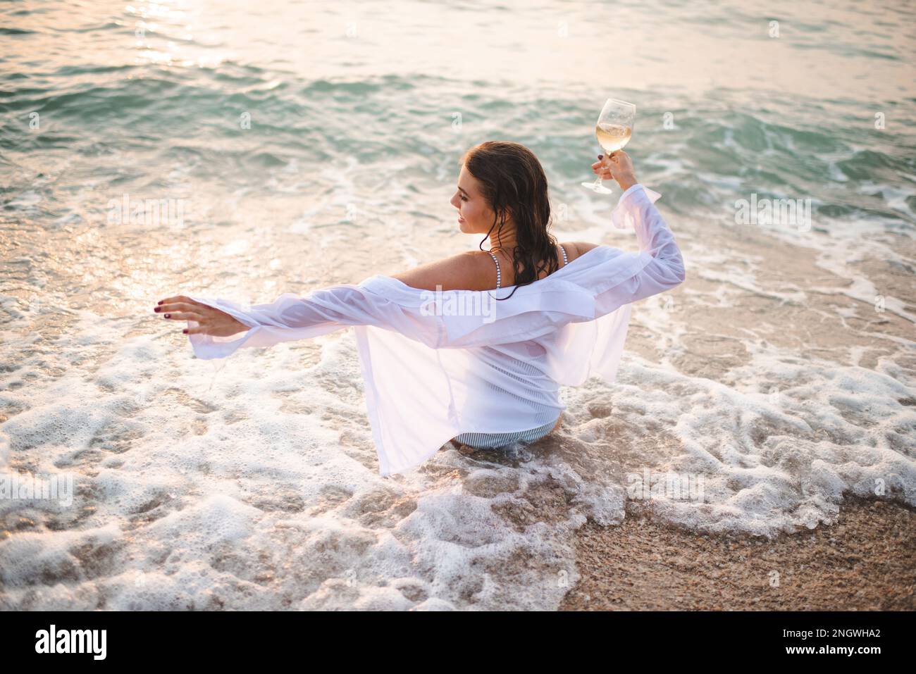 Giovane donna sorridente che tiene un bicchiere di vino bianco seduto in mare sulla costa di sabbia all'aperto al tramonto. Donna felice che indossa una camicia e un costume da bagno bagnati. Summe Foto Stock