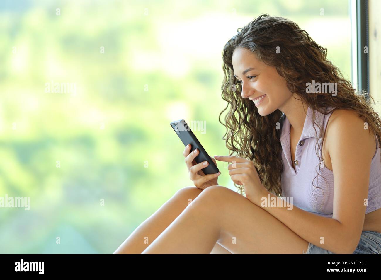 Donna felice che usa lo smartphone vicino a una finestra seduta a casa Foto Stock