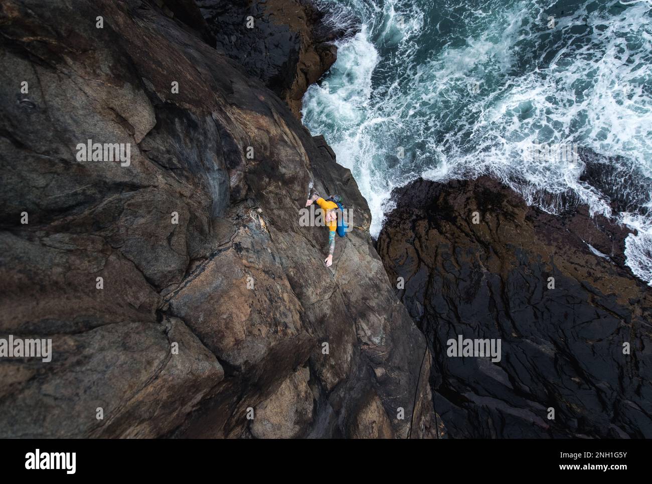 Uomo arrampicata su roccia sulla scogliera del mare con le onde che si schiantano sotto Foto Stock