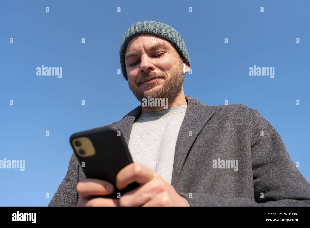 Uomo in cappello e cappotto con il cellulare e sorridente mentre si alza all'aperto contro il cielo blu. Foto Stock