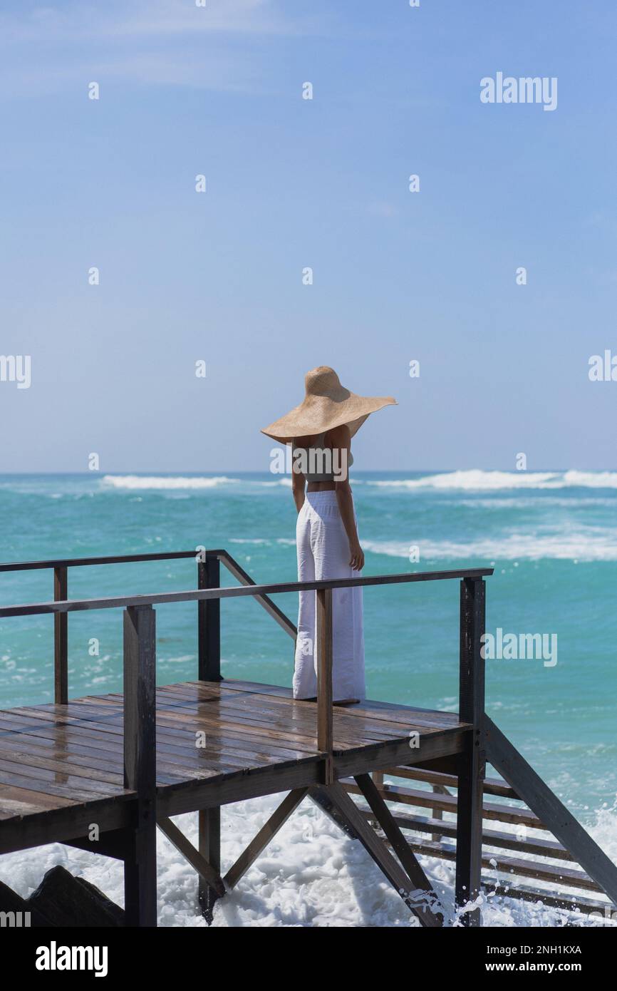 Giovane donna in un cappello di paglia sul molo, le onde dell'oceano schizzare. Foto Stock