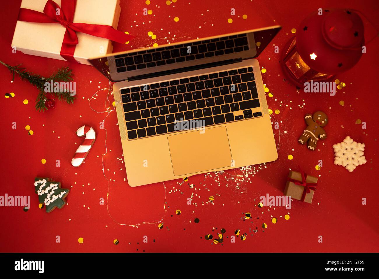 Il computer portatile aperto si trova su un tavolo rosso festivo con glitter e confetti. Ci sono biscotti di Natale e lanterna di Capodanno incandescente. Due regali in kraft Foto Stock