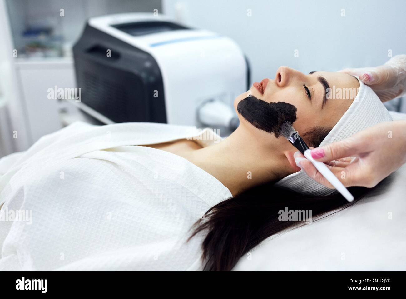 Cosmetologa femminile che applica la maschera nera sul viso di una bella donna per la buccia di carbonio Foto Stock