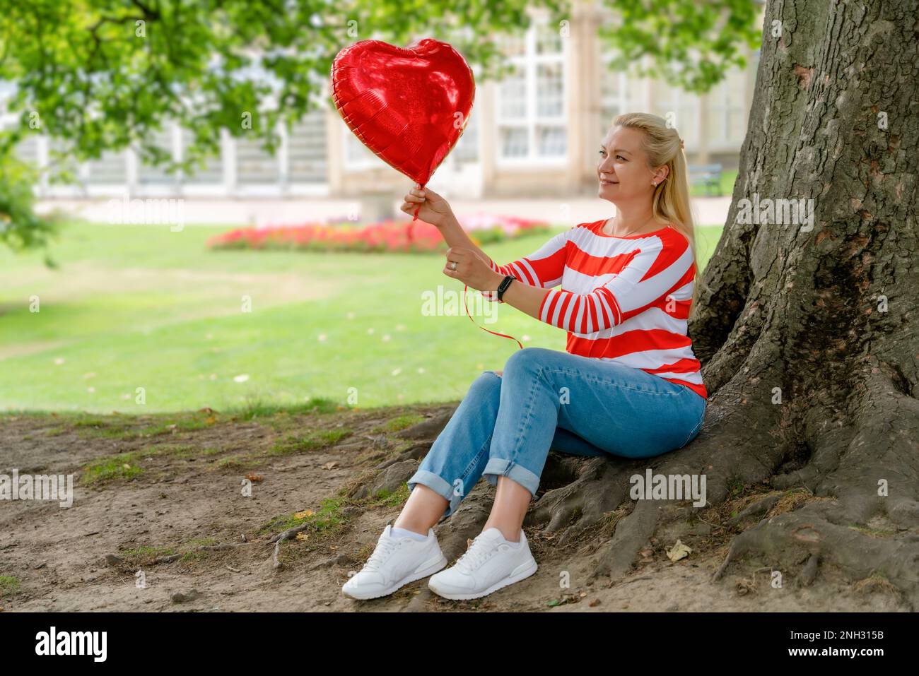 una donna in jeans che tiene un palloncino rosso sotto l'albero nel parco Foto Stock