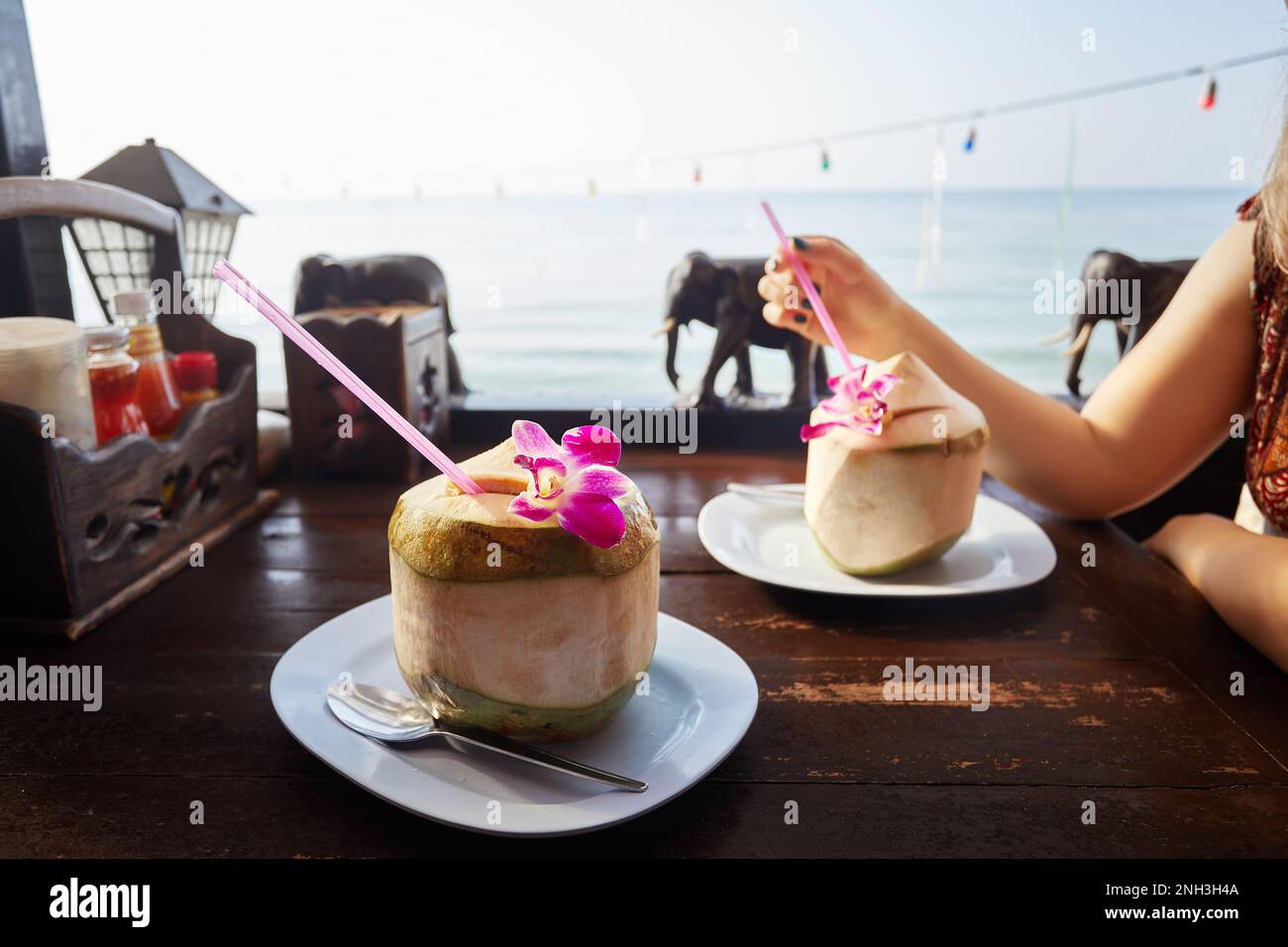 Donna beve dolce cocco fresco con paglia al ristorante con decorazione in legno elefanti sulla spiaggia con vista dell'oceano all'alba. Foto Stock