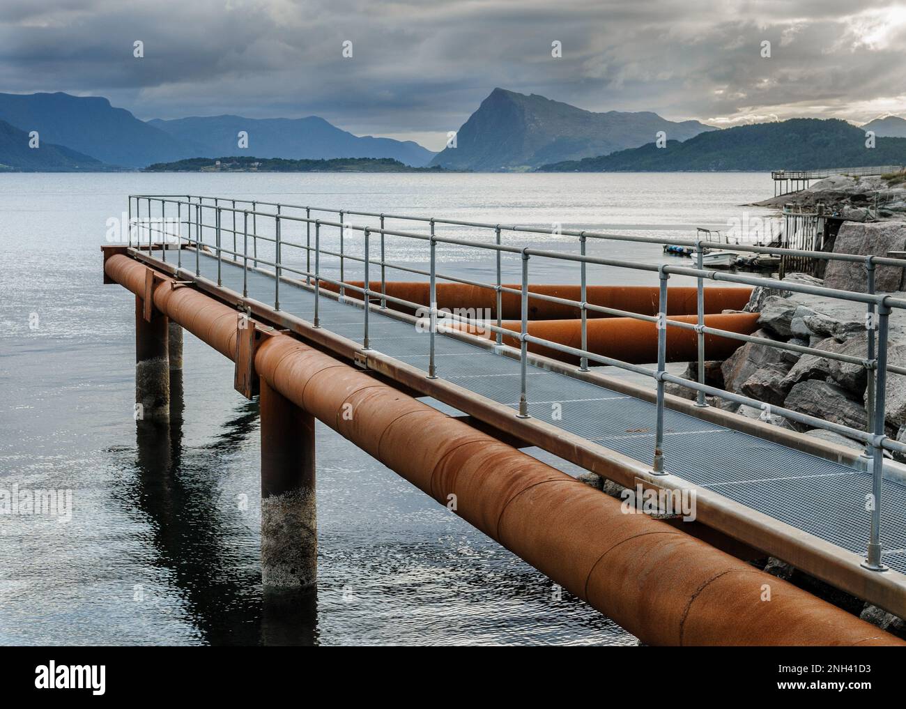 Un oleodotto industriale corre lungo una passerella in legno, che conduce su acque tranquille verso le montagne lontane sotto nuvole grigie. Foto Stock