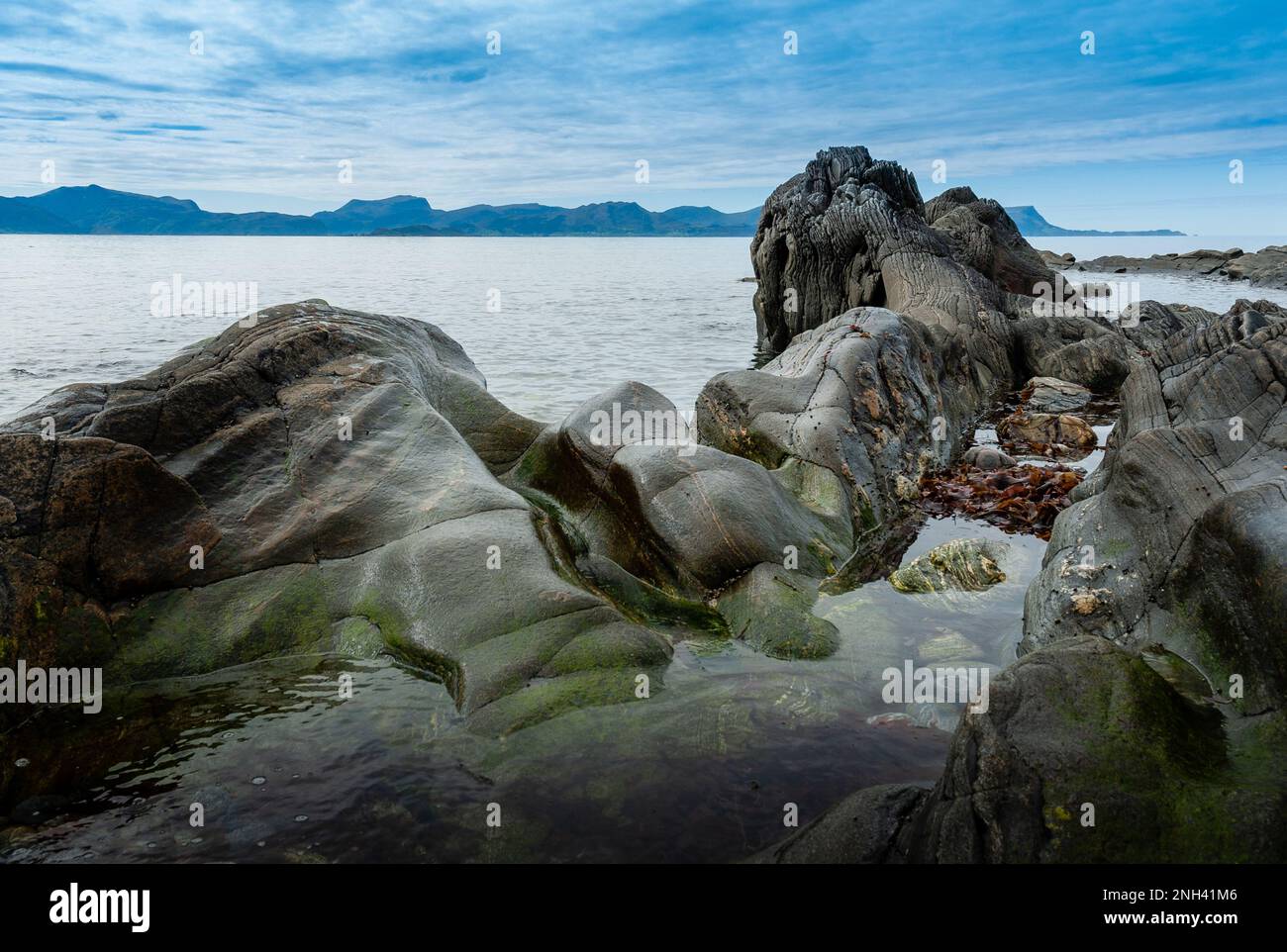 Le onde si infrangono dolcemente contro rocce lisce lungo una costa tranquilla sotto un cielo limpido nel pomeriggio. Foto Stock