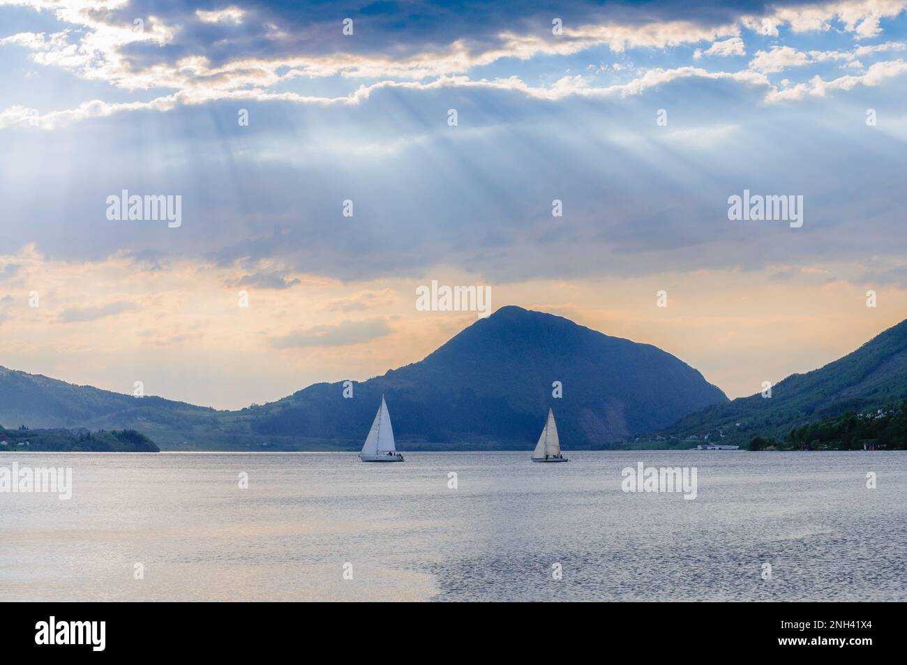 Due barche a vela navigano nelle acque calme di un fiordo, con montagne maestose e luce soffusa dal cielo che creano un'atmosfera tranquilla. Foto Stock