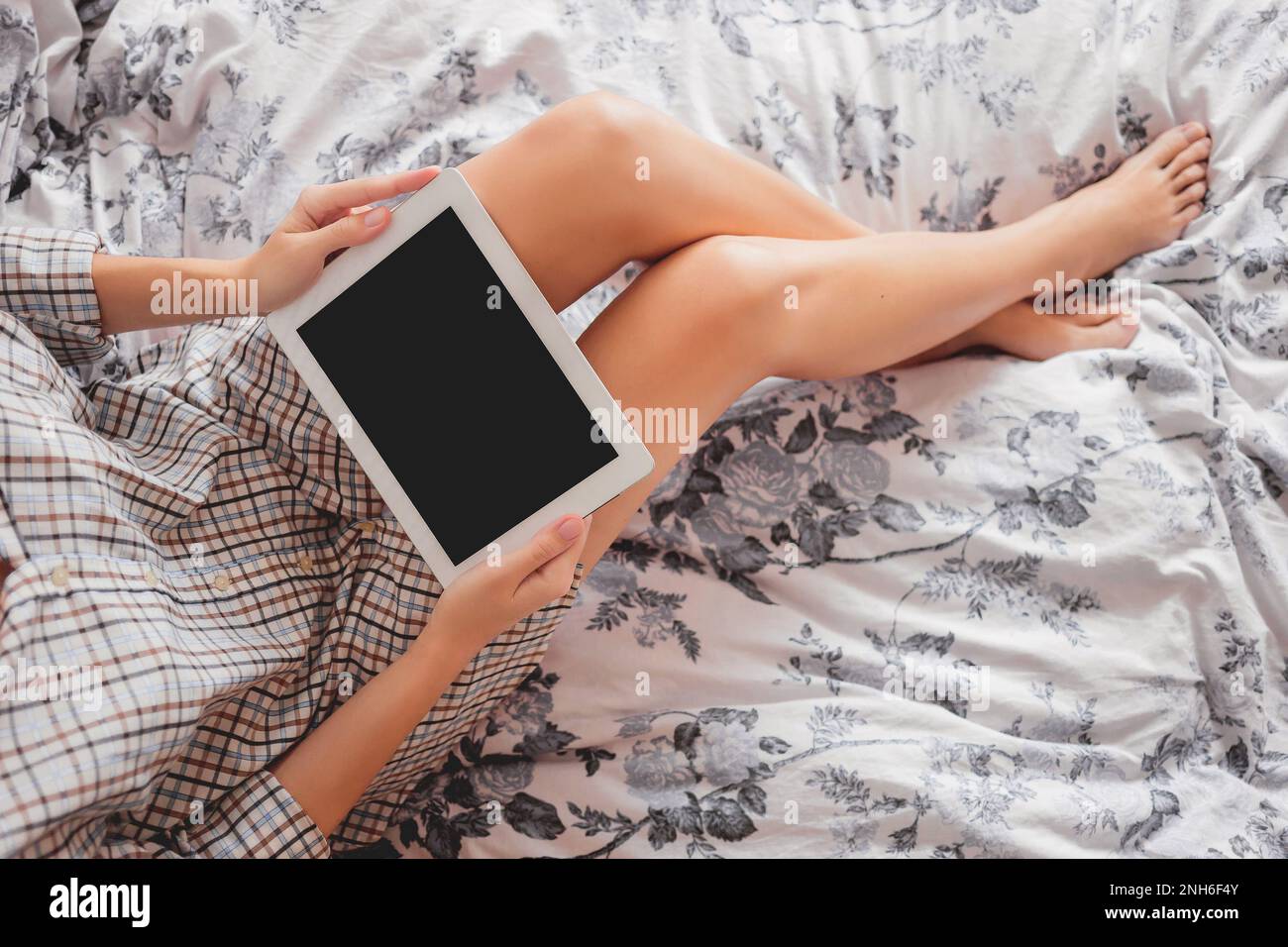 Donna in camicia a scacchi è seduta sul letto con tablet digitale. Accogliente mattina a casa. Foto Stock