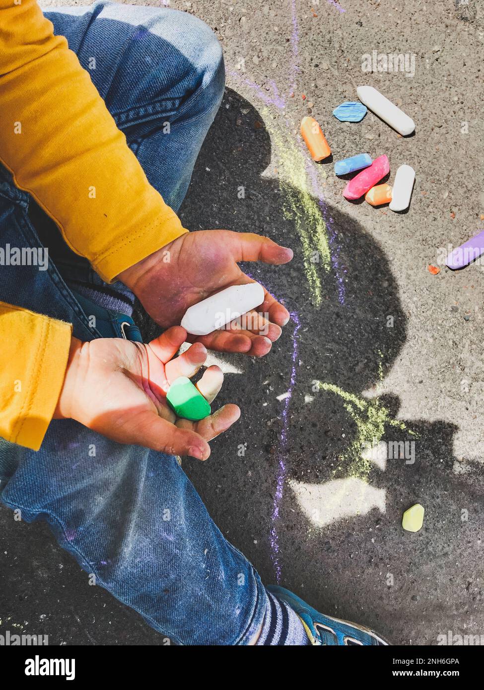 Il bambino in jeans disegna con i pastelli sull'asfalto nelle giornate di sole. Il bambino tiene pastelli colorati. I vestiti dei bambini sono coperti con le macchie colorate Foto Stock