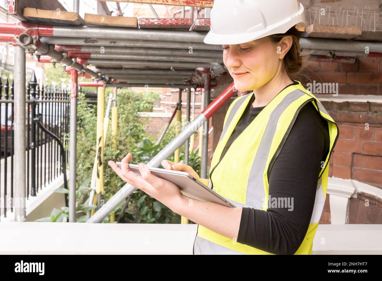 Donna sorridente ingegnere civile prendere appunti su un tablet con una penna elettronica, indossa elmetto e giallo dispositivi di protezione personale, innovazione e. Foto Stock