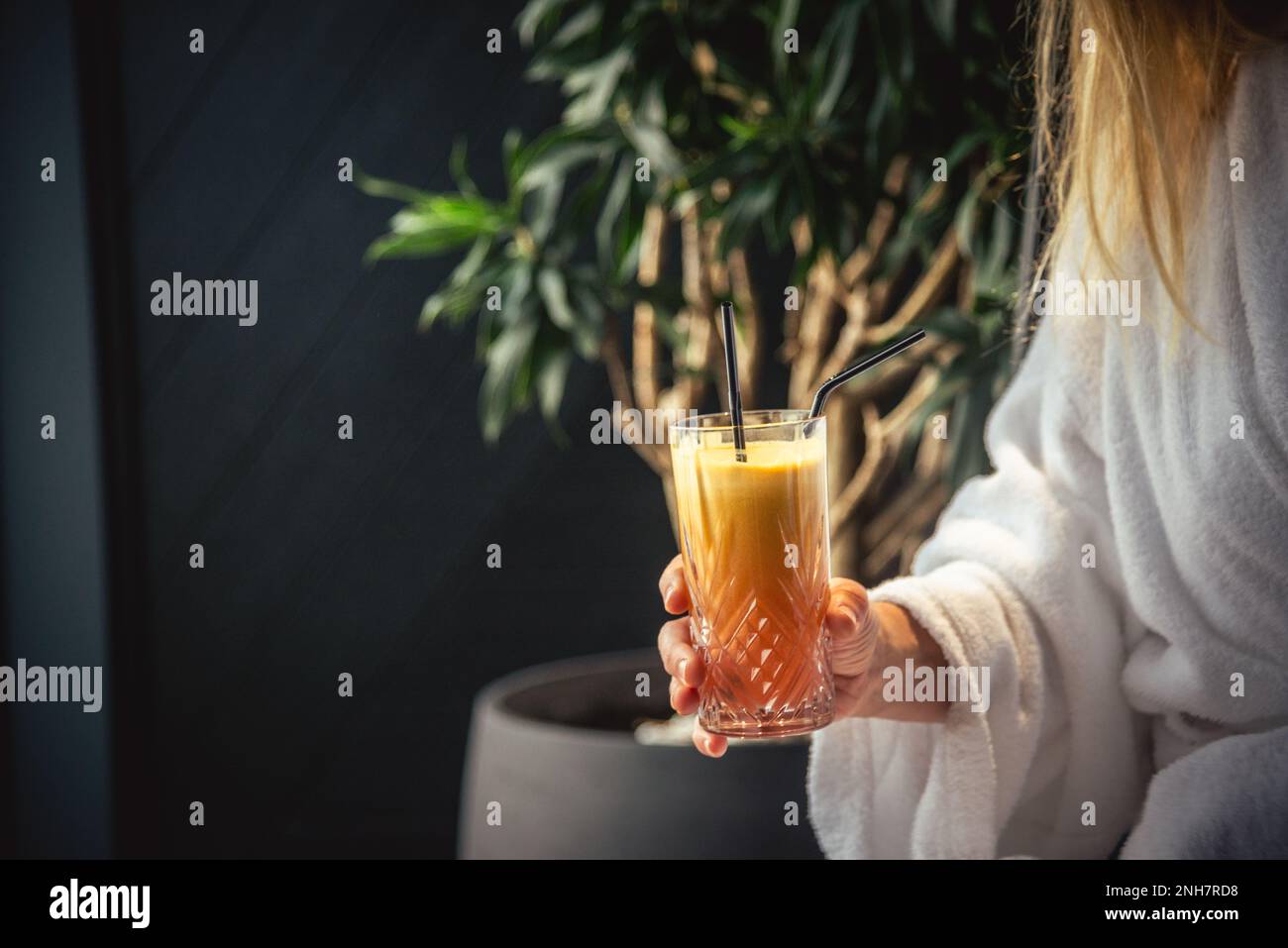 Donna in accappatoio bianco sdraiata sul divano e rilassante con un cocktail arancione a casa vicino alla finestra Foto Stock