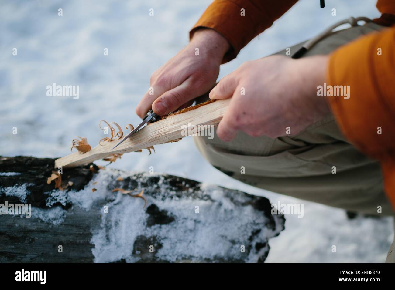 Un uomo prepara lana di legno con un coltello per iniziare un fuoco in una foresta invernale al tramonto. Concetto di sopravvivenza invernale. Foto Stock